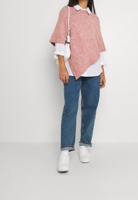 Poncho rose texturé avec un ourlet asymétrique, porté par-dessus une chemise blanche à col. Associé à un jean bleu et des baskets blanches. Petit sac blanc.