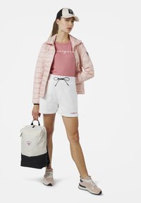 Modellen bär en ljusrosa puffjacka, en rosa t-shirt, vita shorts och grå sneakers, samtidigt som hon håller en beige och svart ryggsäck.