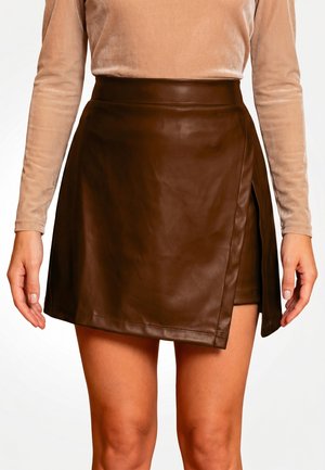Vrouw draagt een donkerbruine leren minirok met een wikkeldetail over blote benen en een beige top met lange mouwen.
