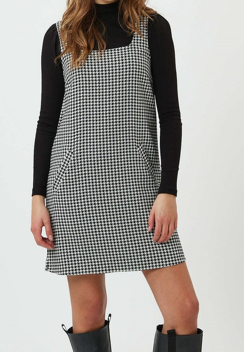 Femme portant un haut noir à manches longues, une robe pied-de-poule noir et blanc avec poches, et des bottes noires mi-mollet, debout devant un fond uni.