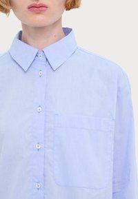 Camicia azzurro chiaro con colletto, dotata di taschino sul petto e una texture sottile. I bottoni sono dello stesso colore del tessuto.