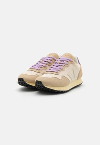 Beige sneakers med overdel i semsket skinn og mesh, lilla lisser og svart gummisåle. Designet inkluderer en hvit logo aksent.