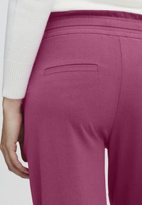 Fitted, high-waisted trousers in vibrant magenta fabric with a smooth texture, featuring a single back pocket and subtle stitch detailing.