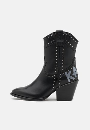 Czarny skórzany botek w stylu western z metalowymi srebrnymi ćwiekami, grubym obcasem, pętelką do zakładania oraz szarym napisem "KARL" po bokach.