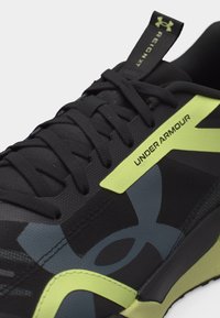 Μαύρο αθλητικό παπούτσι Under Armour Reign XT με κίτρινα σημεία και κορδόνια, με το λογότυπο της μάρκας και ανώτερο μέρος από υφασμάτινο δίχτυ με υφή.