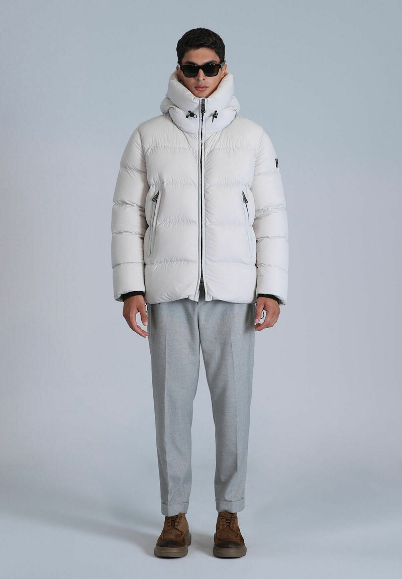 Veste doudoune blanche avec col haut et zips, associée à un pantalon gris clair et des chaussures brunes. Modèle portant des lunettes de soleil.