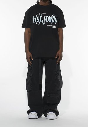 Person trägt schwarze, übergroße Cargohosen, ein schwarzes T-Shirt mit dem Schriftzug „lost youth“ und weiße Turnschuhe, steht vor einem weißen Hintergrund.