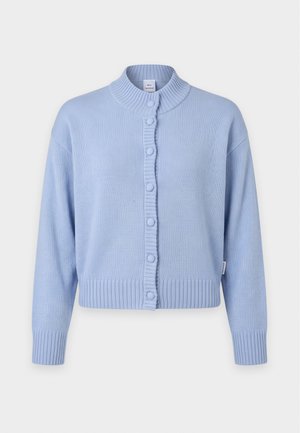 Femme debout portant un cardigan bleu clair sur une blouse blanche, un jean bleu clair avec ceinture noire et des chaussures en cuir marron sur fond gris.