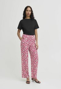 Zwarte korte mouw top met ronde hals, gecombineerd met witte broek met wijde pijpen en een multicolor bloemenpatroon. Model staat met één hand in de zak.