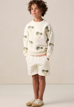 Ensemble composé d'un sweatshirt crème clair et d'un short ornés de motifs de palmiers verts. Texture douce, coupe décontractée et chaussures beiges assorties.