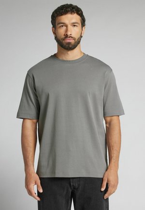 Mand med kort mørkt hår og skæg iført en ensfarvet grå t-shirt med korte ærmer og sorte bukser, stående mod en grå baggrund.