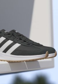 Par de zapatillas negras Adidas con rayas blancas y suelas de goma expuestas sobre un saliente blanco contra un fondo neutro.