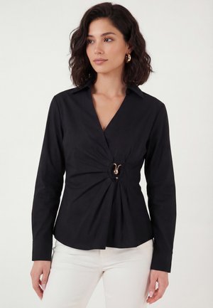 Kvinde med skulderlangt mørkt hår iført guldringøreringe, sort omslagsbluse med central guldlås og hvide bukser, der står mod en ensfarvet baggrund.