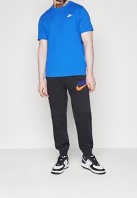 Blå bomullströja med en vit Nike-logotyp, parat med svarta sweatshirtbyxor med en orange Nike-logotyp, tillsammans med svarta och vita sneakers.