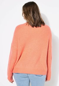 Maglione oversize corallo con texture a coste, maniche ampie e design con spalle scese, abbinato a jeans azzurri chiari.