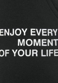 Schwarzer Stoff mit weißem Text, der "GENIEßE JEDEN MOMENT DEINES LEBENS" lautet. Der Text ist fett und befindet sich in der Mitte. Strukturierten Strickhintergrund.