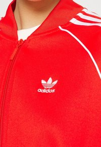 Červená zipová bunda s bílými pruhy a logem, vyrobená z hladké látky. Má vysoký límec a zipové zapínání, s žebrovanými manžetami a lemem.