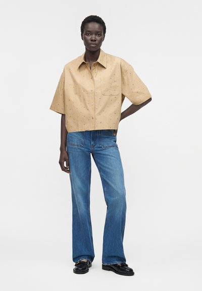 Άτομο που φοράει μπεζ πουκάμισο με κοντά μανίκια και μικρά σχέδια και μπλε τζιν με ίσια πόδια, στέκεται με το ένα χέρι στη μέση.