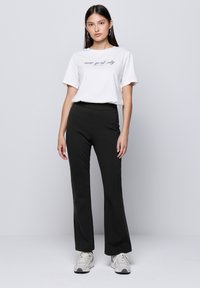 T-shirt blanche à manches courtes avec impression "new york city", associée à un pantalon évasé noir. Le mannequin porte des baskets claires sur un fond uni.