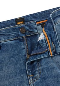 Blå denimjeans delvis neddragna med en synlig metall BOSS-knapp och svart etikett med orange BOSS-text.