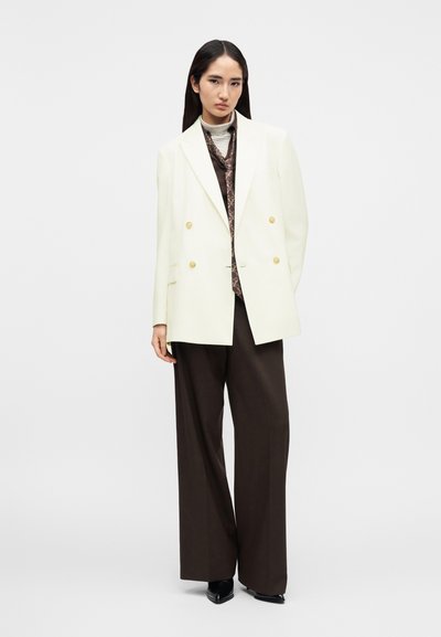 Krém színű, dupla soros arany gombos blézer, barna texturált nyakkendővel és sötét, széles szárú nadrággal párosítva. Elegáns fekete magassarkúkkal kiegészítve.