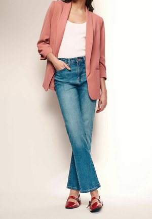Femme portant un blazer rose clair, un haut blanc, un jean bleu et des chaussures plates en cuir verni rouge, debout avec une main dans la poche.