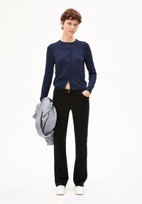 Navy Cardigan mit Knöpfen, kombiniert mit schwarzen Hosen. Trägt eine Jeansjacke, trägt weiße Schuhe. Einfaches, tailliertes Design.