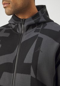 Homme portant un sweat à capuche zippé noir et gris à motifs avec une fermeture éclair à l'avant et un petit logo Jordan sur la poitrine.