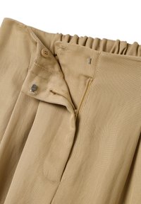 Beige Stoffhose mit gerafftem Bund, ausgestattet mit einem Reißverschluss- und Knopfverschluss sowie seitlichen Taschen. Die Textur ist gerippt.