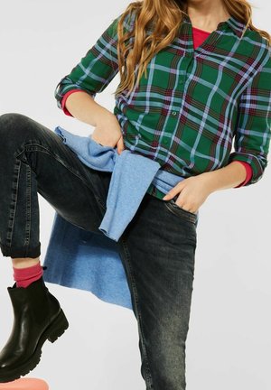 Groene en blauwe geruite overhemd, zwarte jeans, blauwe trui om de taille geknoopt, zwarte enkellaarzen, met zichtbare roze sokken.