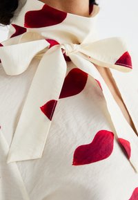 Tissu de couleur crème avec de grands motifs de cœurs rouges, doté d'un col en nœud papillon et d'une texture lisse. Le tissu a un léger éclat et une douceur au drapé.