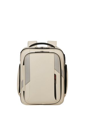 Beiger rechteckiger Rucksack mit schwarzen Reißverschlüssen, gepolsterten Trägern, Fronttasche mit Mesh-Details und einem oberen Tragegriff.