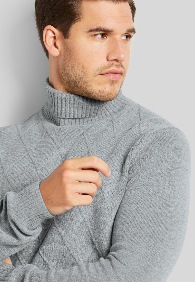 Grauer Pullover mit geripptem Rollkragen, ausgestattet mit einem Diamantmuster, strukturierter Stoff und eng anliegenden Ärmeln. Hand in der Nähe des Kragens positioniert.