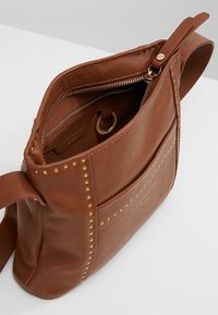 Hnědá kožená crossbody taška se zdobením, s kapsou na zip a měkkou texturou. Obsahuje odnímatelný kroužek na klíče uvnitř.