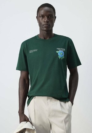 Hombre con camiseta verde oscuro con texto blanco y un gráfico de una flor azul, pantalones beige, de pie frente a un fondo claro y liso.