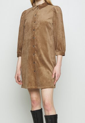 Femme portant une robe en daim marron boutonnée avec des manches ballon trois-quarts et des bottes noires montantes jusqu'aux genoux, debout devant un fond uni.