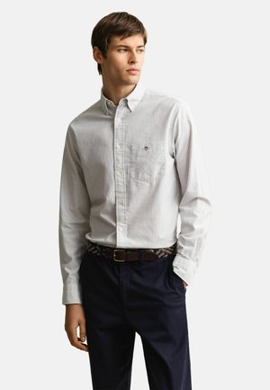 Giovane uomo che indossa una camicia a righe grigio chiaro con bottoni, infilata in pantaloni blu scuro con una cintura intrecciata marrone, che guarda verso destra.