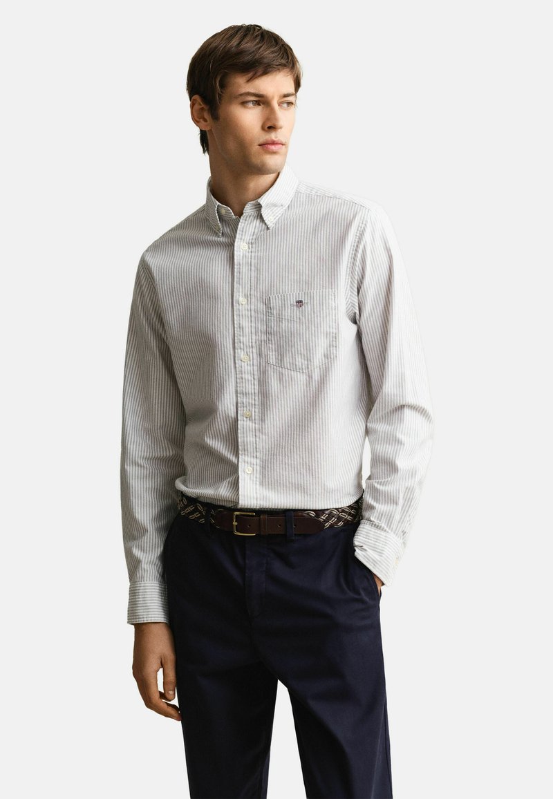 Junger Mann, der ein hellgraues, gestreiftes Hemd mit Knöpfen trägt, das in dunkelblaue Hosen mit einem braunen geflochtenen Gürtel gesteckt ist, und nach rechts blickt.