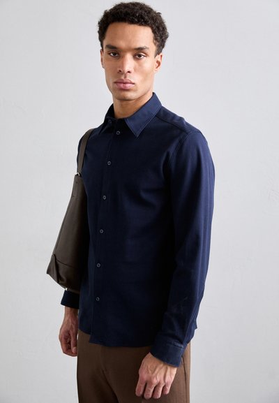 Camisa de botones de manga larga en azul marino con un cuello con solapa, diseño ajustado y un bolsillo en el pecho. Combinada con un bolso de hombro marrón.