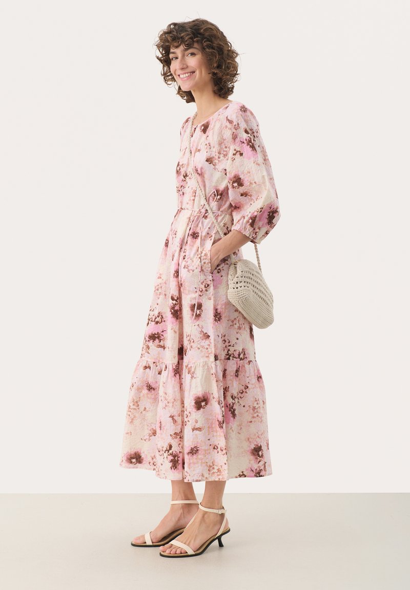 Robe midi florale en rose et crème avec des manches bouffantes, une ceinture à la taille, un ourlet évasé et des volants superposés ; associée à un sac en crochet beige et des talons.