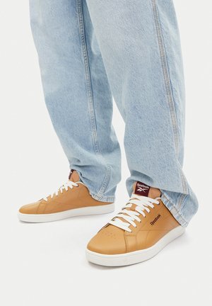 Hellblaue Jeans, die beige Reebok-Sneaker mit weißen Schnürsenkeln und Sohlen bedecken, stehend auf weißem Hintergrund.