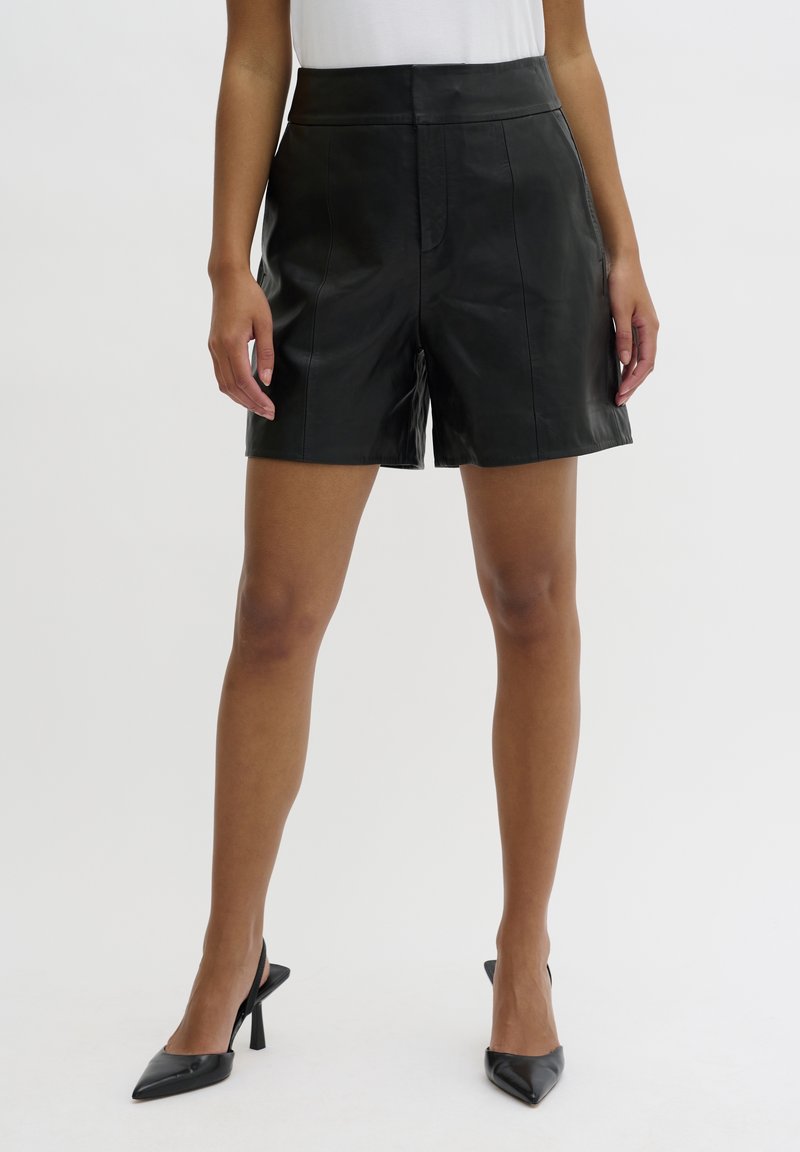 Femme portant un short en cuir noir taille haute associé à des escarpins slingback noirs à bout pointu sur un fond uni.
