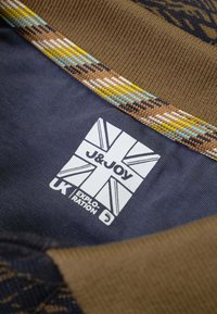 Bruine stoffen kraag met een textuurontwerp; onderaan bevindt zich een marineblauwe stof met een wit label waarop "J&JOY" en een Union Jack-graphic staat.