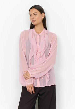 Femme aux longs cheveux foncés porte un chemisier léger rose clair transparent à volants, associé à un pantalon foncé, debout devant un fond blanc.