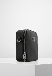 Borsa nera con zip di forma quadrata, superficie texturizzata, dettaglio con logo e minuteria in argento. Presenta un anello laterale per il fissaggio.