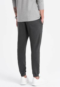 Pantalones de chándal grises con puños elásticos, dos bolsillos traseros y una textura suave. Combinados con zapatillas blancas, vistos desde atrás.