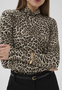 Leopardenmuster Rollkragenoberteil in Beige und Braun, mit langen Ärmeln und einem goldenen Armband-Akzent, kombiniert mit schwarzer Hose und Gürtel.