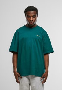 Tricou oversize din bumbac turquoise cu mâneci scurte, având un mic logo brodat cu fir aurit pe pieptul stâng. Textură netedă.