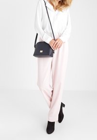 Borsa a tracolla in pelle nera con forma curva, hardware dorato e finitura testurizzata, abbinata a pantaloni rosa chiaro e una camicia bianca.
