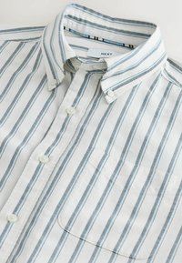 Hvid skjorte med knapper foran med lodrette grå striber, en brystlomme og en krave med knapper, lagt fladt på en lys overflade.